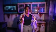 A person appears surprised or shocked while standing in a dimly lit, disheveled room with smeared blood, conveying a sense of horror or suspense.