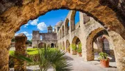 The image showcases the historic stone arches and architecture of an ancient mission against a backdrop of a bright blue sky with fluffy clouds.