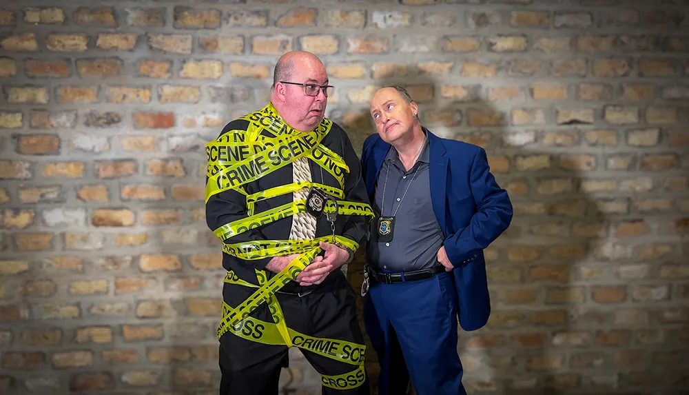 A person wrapped in yellow crime scene tape stands next to another individual posing as a police officer against a brick wall backdrop creating a humorous scene