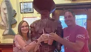 A man and a woman are smiling and toasting with drinks next to a large wooden carving of what appears to be a pirate, in a restaurant adorned with a fish wall mount and nautical photographs.